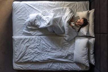 Genç bir kadın yatağa uzanıyor ve akıllı telefon kullanıyor. Uyan ve rahat bir yatak odasında cep telefonunda internete göz at. Otelde dinlenen bir kadın, tembel bir sabah. Yukarıdan görüntüle.