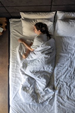 Genç esmer bir kadın pijamalarıyla battaniyenin altında yatıyor, yumuşak bir yastıkta uyuyor, tatlı rüyaların tadını çıkarıyor, yatakta dinleniyor. Üst görünüm.