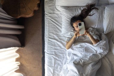Genç bir kadın yatağa uzanıyor ve akıllı telefon kullanıyor. Sabahları yatakta telefonlu bir kadın. Otelde dinlenen bir kadın, tembel bir sabah. Yukarıdan görüntüle.