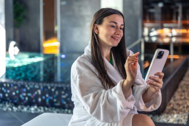 Beyaz cüppeli çekici genç bir kadın spa kompleksinde dinleniyor. Bir kadın saunada akıllı telefon kullanıyor. Boşluğu kopyala.