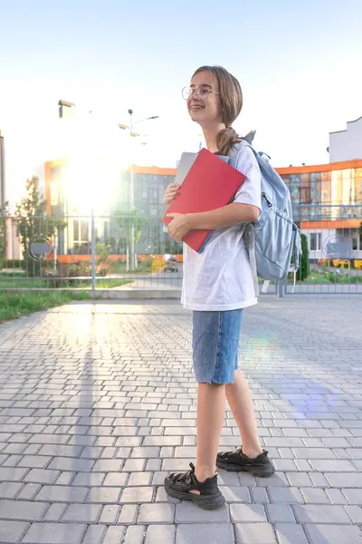 Ellerinde defterleri ve omuzlarında sırt çantası olan bir kız öğrenci. Günbatımı okulun arka planında. Güzel gülüşlü bir kız beyaz bir tişört giyiyor. Yüksek kalite fotoğraf