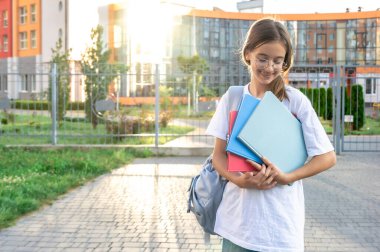 Ellerinde defterleri ve omuzlarında sırt çantası olan bir kız öğrenci. Günbatımı okulun arka planında. Güzel gülüşlü bir kız beyaz bir tişört giyiyor. Yüksek kalite fotoğraf