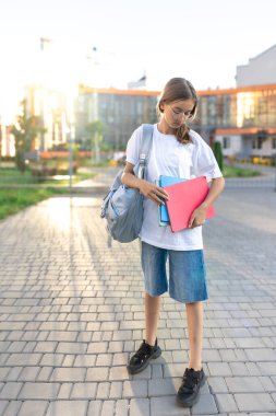 Ellerinde defterleri ve omuzlarında sırt çantası olan bir kız öğrenci. Günbatımı okulun arka planında. Güzel gülüşlü bir kız beyaz bir tişört giyiyor. Yüksek kalite fotoğraf