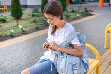 Öğrenci okulun yanında kalemlerle bir bankta oturuyor. Sırt çantalı bir kız. Liseli kız okul malzemeleriyle bir bankta oturuyor, beyaz bir tişört giyiyor. Yüksek kalite fotoğraf