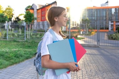 Ellerinde defterleri ve omuzlarında sırt çantası olan bir kız öğrenci. Günbatımı okulun arka planında. Güzel gülüşlü bir kız beyaz bir tişört giyiyor. Yüksek kalite fotoğraf