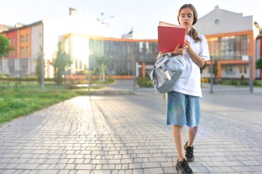 Ellerinde defterleri ve omuzlarında sırt çantası olan bir kız öğrenci. Günbatımı okulun arka planında. Güzel gülüşlü bir kız beyaz bir tişört giyiyor. Yüksek kalite fotoğraf