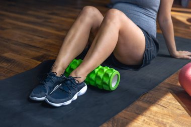 Hamile kadın spor salonunda masaj rulosuyla egzersiz yapıyor, paspasın üzerinde oturuyor. Sağlıklı spor yaşam tarzı.