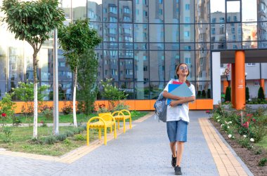 Ellerinde defterleri ve omuzlarında sırt çantası olan bir kız öğrenci. Günbatımı okulun arka planında. Güzel gülüşlü bir kız beyaz bir tişört giyiyor. Yüksek kalite fotoğraf