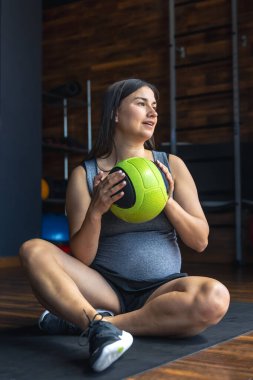 Spor salonundaki hamile bir kadın elinde topla bir paspasın üzerinde oturuyor. Spor yaşam tarzı konsepti. Yüksek kalite fotoğraf