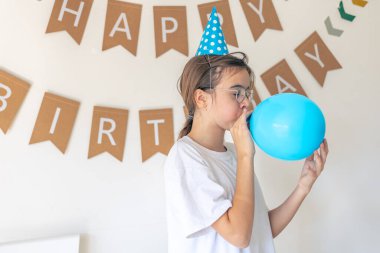 Neşeli bir kız doğum gününde balon şişirir. Yüksek kalite fotoğraf. Şenlik şapkalı bir kız. Kız mavi bir top şişiriyor. Doğum günü konsepti. Metin için boşluk.