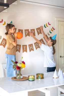 İki kız kardeş duvara doğum günü yazısını astı. Doğum günü konsepti. Yüksek kalite fotoğraf. Şenlik şapkalı kızlar. Doğum günü hazırlığı..