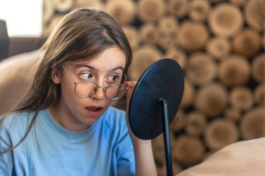 Gözlüklü güzel bir kız aynada kendine bakıyor. Sabah rutini. Yüksek kalite fotoğraf. Kız evdeki aynada kendine bakıyor. Metin için boşluk.