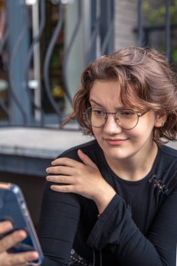 Telefonlu genç bir kadın. Sürpriz bir yakınlaşma hissi. Yüksek kalite fotoğraf. Gözlüklü, telefonu olan bir kız. Video çağrısı sohbeti veya arkadaşlarla yazışma.