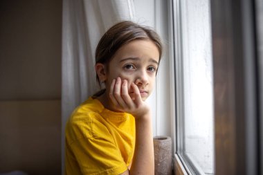 Kasvetli ve üzgün bir kız pencereden dışarı bakar. Duygular konsepti. Yüksek kalite fotoğraf. Duygusal hüzünlü yakın çekim. Kızın üzücü düşünceleri var. Okul çağındaki üzgün bir kızın portresi..