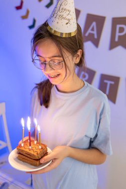 Mutlu bir kız elinde mumlarla şenlikli bir pasta tutuyor. Doğum günü konsepti. Yüksek kalite fotoğraf. Gözlüklü ve neşeli şapkalı bir kız..