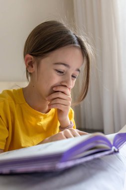 İlgili ve mutlu bir kız bir kitap okuyor. Duygu konsepti. Yüksek kalite fotoğraf. Okul çağındaki bir kız ilgilenir ve odaklanır. Evde ödev yapıyorum..