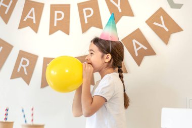 Doğum gününde balon şişiren tatlı bir kız. Evde doğum günü partisi. Yüksek kalite fotoğraf. Sarı balon. Duvardaki yazı doğum günün kutlu olsun. Şapkalı bir kız..