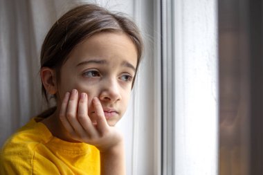 Kasvetli ve üzgün bir kız pencereden dışarı bakar. Duygular konsepti. Yüksek kalite fotoğraf. Duygusal hüzünlü yakın çekim. Kızın üzücü düşünceleri var. Okul çağındaki üzgün bir kızın portresi. Metin için boşluk.