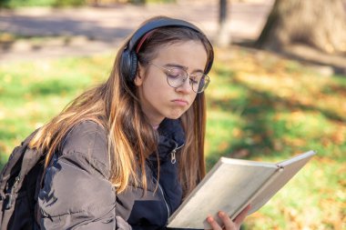 Üzgün bir kız kulaklıkla müzik dinler ve kitap okur. Yüksek kalite fotoğraf. Duygular konsepti. Kız bir kitap okuyor ve tek başına yürürken müzik dinliyor..