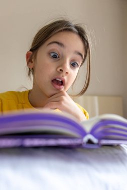 İlgili ve mutlu bir kız bir kitap okuyor. Duygu konsepti. Yüksek kalite fotoğraf. Okul çağındaki bir kız ilgilenir ve odaklanır. Evde ödev yapıyorum..