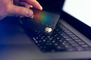 Ukrayna, Vinnytsia - 11 Aralık 2024: Bir el bilgisayar arka planında bir Mastercard banka kartını tutuyor. Modern tasarım, yansıtıcı yüzey, detaylı dokular, gerçekçi parlak ışık. Kapat..