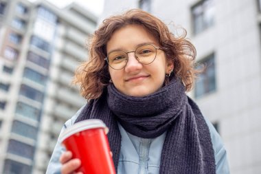 Şehirde elinde kahve olan öğrenci bir kız. Kameraya bakıyorum. Yüksek kalite fotoğraf. Yakın plan. Gözlüklü ve siyah atkılı kız. Büyük bir şehirde öğrenci hayatı.
