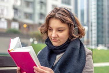 Şehirde kitap okuyan mutlu bir genç kız. Kız kitap okuyor ve müzik dinliyor. Yüksek kalite fotoğraf. Kız bankta otururken kitap okuyor..