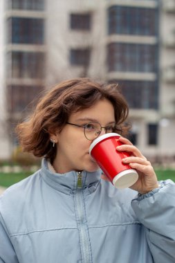 Güzel genç bir kadın büyük bir şehirde kahve içiyor. Şehirdeki kadın. Yüksek kalite fotoğraf. Bir kadın sokakta yürüyor ve elinde kahvenin tadını çıkarıyor. Gözlüklü kız. Metin için boşluk.