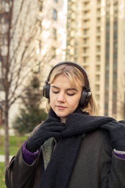 Güzel genç bir kadın kulaklıkla müzik dinliyor ve şehirde dolaşıyor. Yüksek kalite fotoğraf. Sonbahar sezonu, ceket ve fularlı kız. Mesaj için yer var. Müzikten hoşlanan kadın.