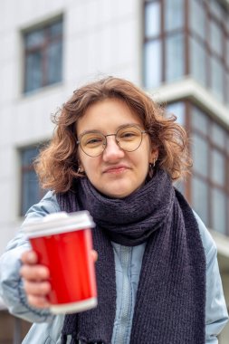 Şehirde elinde kahve olan öğrenci bir kız. Kameraya bakıyorum. Yüksek kalite fotoğraf. Yakın plan. Gözlüklü ve siyah atkılı kız. Büyük bir şehirde öğrenci hayatı.