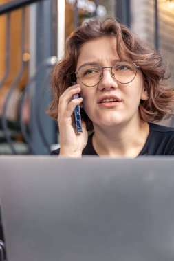 Busy young woman talking on the phone and working on a laptop. High quality photo. A girl in glasses is talking on the phone and a laptop is next to her. Emotion concept. clipart