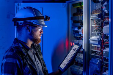Elektrikçi, sigortalı santralde tablet kullanarak çalışmaya odaklanmış. Arka planda acil durum ışıkları ve karanlık odada el feneri olan özel kıyafetli genç bir elektrik mühendisi..