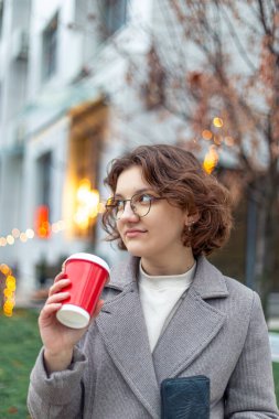 Genç bir kız öğrenci elinde bir kahveyle şehirde yürüyor. Yüksek kalite fotoğraf. Gözlüklü, mavi ceketli ve siyah atkılı kız..