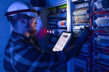 Elektrikçi, sigortalı santralde tablet kullanarak çalışmaya odaklanmış. Arka planda acil durum ışıkları ve karanlık odada el feneri olan özel kıyafetli genç bir elektrik mühendisi..