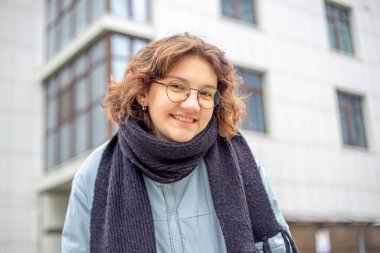 Şehirde mutlu genç bir kadın. Şehirde yürüyordum. Yüksek kalite fotoğraf. Yüksek binaların yanında gözlüklü mutlu kız. Mavi ceketli ve siyah atkılı kız kameraya bakıyor..