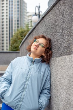 Büyük bir şehirde genç bir kadın. Şehrin tadını çıkaran bir kız. Mesaj için yer var. Yüksek kalite fotoğraf. Gözlüklü ve mavi ceketli kız. Şehirde mutlu bir kız. Kısa saçlı. Arka planda yüksek binalar var.
