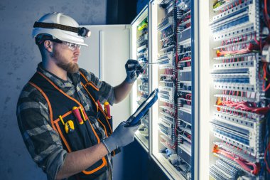 Elektrikçi, sigortalı santralde tablet kullanarak çalışmaya odaklanmış. Arka planda acil durum ışıkları ve karanlık odada el feneri olan özel kıyafetli genç bir elektrik mühendisi..