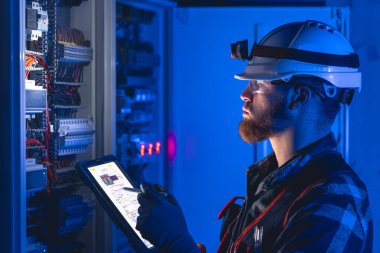 Elektrikçi, sigortalı santralde tablet kullanarak çalışmaya odaklanmış. Arka planda acil durum ışıkları ve karanlık odada el feneri olan özel kıyafetli genç bir elektrik mühendisi..