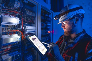 Elektrikçi, sigortalı santralde tablet kullanarak çalışmaya odaklanmış. Arka planda acil durum ışıkları ve karanlık odada el feneri olan özel kıyafetli genç bir elektrik mühendisi..