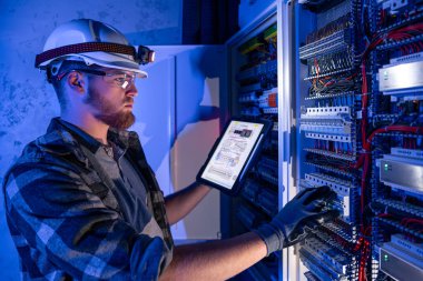 Elektrikçi, sigortalı santralde tablet kullanarak çalışmaya odaklanmış. Arka planda acil durum ışıkları ve karanlık odada el feneri olan özel kıyafetli genç bir elektrik mühendisi..