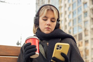 Şehirde telefonu olan çekici bir kadın. Açık havada müzik dinlemek. Yüksek kalite fotoğraf. Bir kız kahve dükkanından güzel kokulu kahve içer, müzik dinler ve telefonunu kullanır..