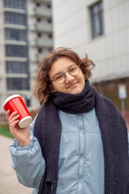 Şehirde elinde kahve olan öğrenci bir kız. Kameraya bakıyorum. Yüksek kalite fotoğraf. Yakın plan. Gözlüklü ve siyah atkılı kız. Büyük bir şehirde öğrenci hayatı.