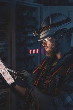 Elektrikçi, sigortalı santralde tablet kullanarak çalışmaya odaklanmış. Arka planda acil durum ışıkları ve karanlık odada el feneri olan özel kıyafetli genç bir elektrik mühendisi..