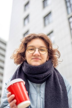Şehirde elinde kahve olan öğrenci bir kız. Kameraya bakıyorum. Yüksek kalite fotoğraf. Yakın plan. Mesaj için yer var. Gözlüklü ve siyah atkılı kız..