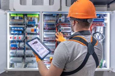 Tulumlu bir erkek elektrikçi, sigortalı ve tablet kullanan bir santralde çalışmaya odaklanmış. Yüksek kalite fotoğraf. Elinde tablet olan bir elektrikçi santralde çalışıyor. Boşluğu kopyala.