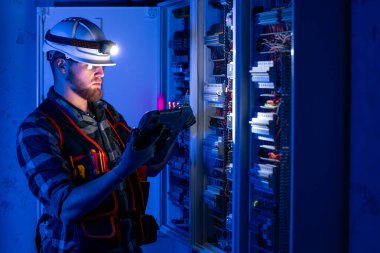 Elektrikçi, sigortalı santralde tablet kullanarak çalışmaya odaklanmış. Arka planda acil durum ışıkları ve karanlık odada el feneri olan özel kıyafetli genç bir elektrik mühendisi..