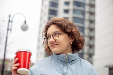 Genç bir kız öğrenci elinde bir kahveyle şehirde yürüyor. Yüksek kalite fotoğraf. Gözlüklü, mavi ceketli ve siyah atkılı kız..