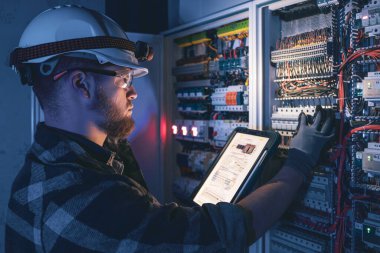 Elektrikçi, sigortalı santralde tablet kullanarak çalışmaya odaklanmış. Arka planda acil durum ışıkları ve karanlık odada el feneri olan özel kıyafetli genç bir elektrik mühendisi..