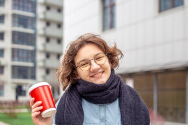 Şehirde elinde kahve olan öğrenci bir kız. Kameraya bakıyorum. Yüksek kalite fotoğraf. Yakın plan. Gözlüklü ve siyah atkılı kız. Büyük bir şehirde öğrenci hayatı.