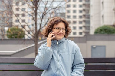 Şehirde genç bir kadın telefonda konuşuyor. Soğuk havada telefonda konuşan bir kız. Yüksek kalite fotoğraf. Gözlüklü, siyah eşarplı ve mavi ceketli bir kız. İş kadını telefonda konuşuyor..